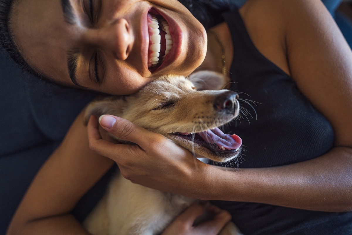 Foto colorida. Mulher negra sorridente abraça um cachorrinho amarelo - Metrópoles