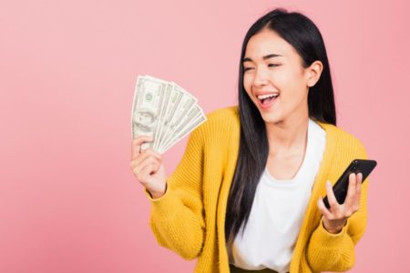 Mulher segurando dinheiro nas mãos e sorrindo com o celular na outra mão de blusa amarela - Metrópoles