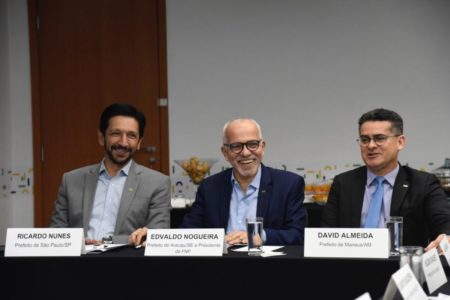 Imagem colorida mostra mesa de reunião com Ricardo Nunes, homem branco de cabelo e barba pretos, terno cinza e camisa azul, ao lado de dois homens - Metrópoles