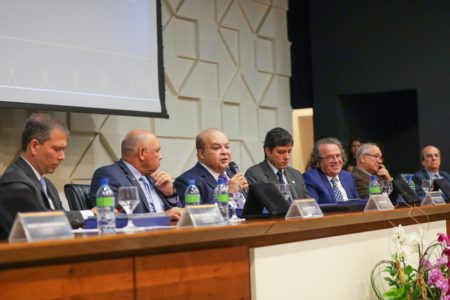 Foto colorida de sete homens, vestidos com ternos, sentados à tribuna, à frente de microfones, em evento do setor produtivo do Distrito Federal. Ao centro, o governador Ibaneis Rocha fala ao microfone