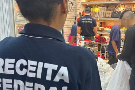 Agente da Receita Federal olha para comércio; pessoas seguram sacolas grandes - Metrópoles