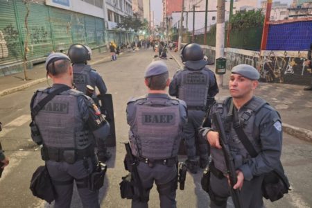 Foto colorida de cinco policiais militares armados em operação na cracolândia com usuários de drogas ao fundo - metrópoles