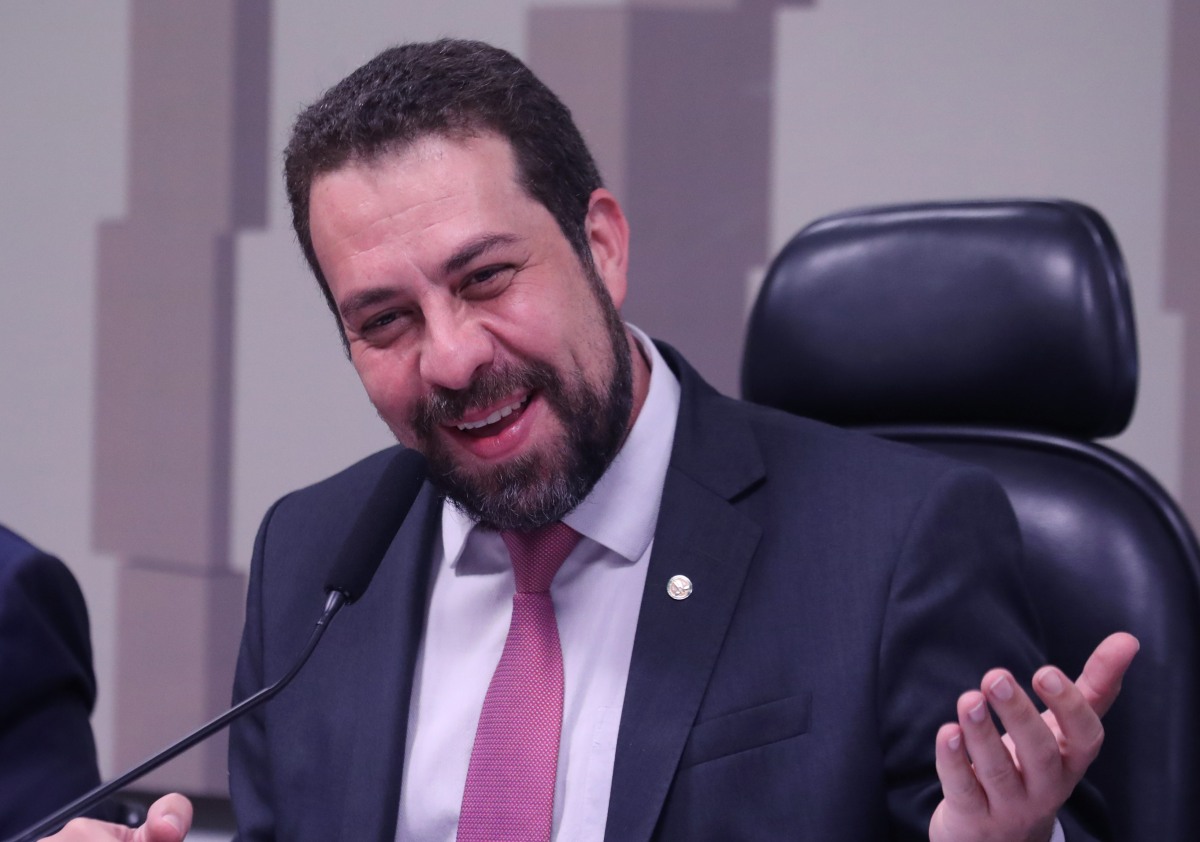 Imagem colorida de Guilherme Boulos, homem branco de cabelo e barba castanhos, de terno azul e gravata rosa, sentado em uma cadeira de couro, de frente a um microfone, sorrindo - Metrópoles