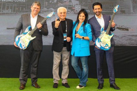 Imagem colorida mostra quatro pessoas em pé, em um piso de grama sintética com um painel publicitário ao fundo. Os homens das duas pontas, Tarcísio de Freitas e Ricardo Nunes, seguram guitarras azuis. Ambos estão vestidos de palotó - Metrópoles