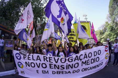 Imagem colorida mostra grupo de estudantes segurando uma faixa branca com letras pretas com a frase "Pela revogação do novo ensino médio e em defesa do orçamento". As pessoas ao fundo seguram várias bandeiras de entidades estudantis - Metrópoles