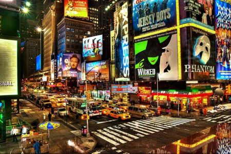 Imagem colorida da Broadway, rua movimentada com uma série de paineis de propaganda cobrindo a fachada de prédios - Metrópoles