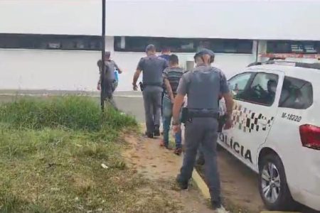 Imagem colorida mostra três policiais militares conduzindo bandidos que aplicaram o golpe do Tinder em um rapaz na zona norte de São Paulo. Um dos bandidos veste camiseta azul e outro veste uma camiseta listrada horizontalmente nas cortes cinza e preta. Eles e os policiais estão de costas ao lado de uma viatura da PM - Metrópoles