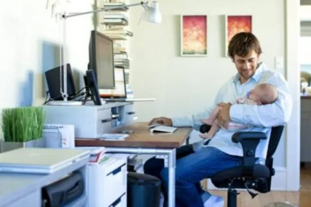 Imagem de homem, em sua casa, sentado na cadeira do escritório, segurando o filho, um bebê, enquanto trabalha. O homem veste uma camisa branca e calça jeans azul e olha para a criança enquanto mexe no mouse - Metrópoles