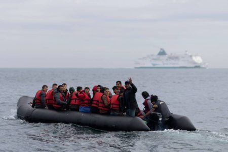 imagem colorida mostra barco em alto mar cheio de imigrantes com colete salva vidas, com navio ao fundo - Metrópoles