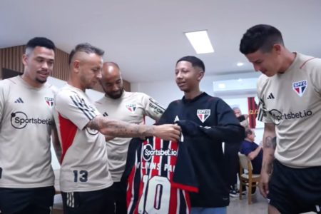 Foto colorida mostra garoto no centro, de casaco preto do São Paulo, recebendo uma camisa das mãos do jogador Rafinha, que está ao seu lado. Os atletas James Rodrigues, Lucas Moura e Luciano também aparecem.