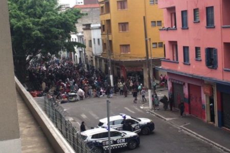 Imagem colorida mostra cruzamento de ruas da Cracolândia, no centro de SP, com uma multidão de usuários de crack ao fundo e dois veículos policiais perto - Metrópoles