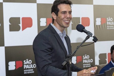 Imagem colorida mostra o secretário estadual da Educação, Renato Feder, homem branco, de cabelo castanho claro e terno cinza. Ele está sorrindo enquanto fala em um microfone, em um púlpito, com um banner com logotipos do governo de SP ao fundo - Metrópoles