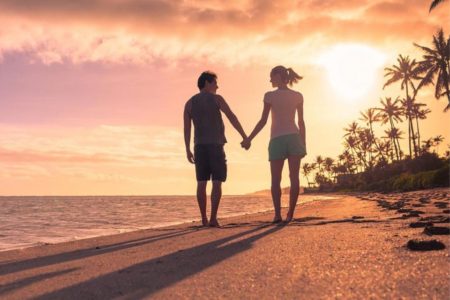 Casal andando de mãos dadas na praia ao por do sol - Metrópoles