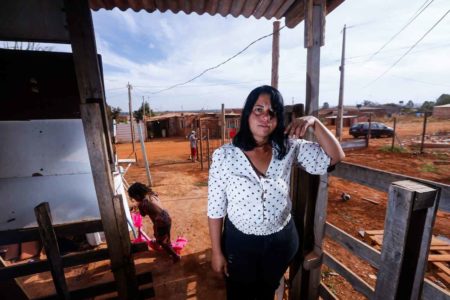 Mulher apoia braço em grade de madeira, criança de 2 anos brinca com patinete rosa ao lado