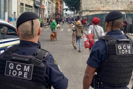 Imagem colorida mostra dois agentes da CGM/IOPE de costas; ao fundo, usuários de drogas transitam em rua estreita - Metrópoles