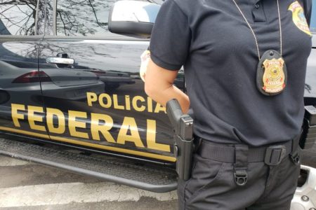 Dino PF Imagem colorida mostra parte de viatura da Polícia Federal; um agente está com braços cruzados e distintivo à mostra - Metrópoles