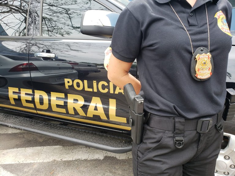 Dino PF Imagem colorida mostra parte de viatura da Polícia Federal; um agente está com braços cruzados e distintivo à mostra - Metrópoles