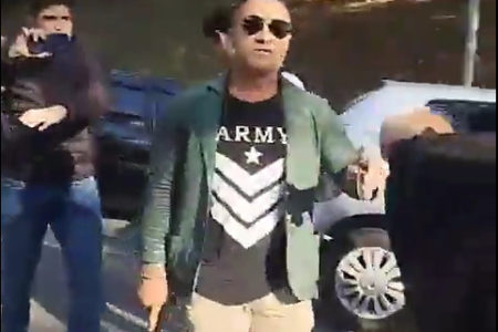 Imagem colorida de homem com camiseta preta, blusa azul e óculos escuros segura revólver apontado para baixo, euquanto sinaliza com outro braço para pessoas se afastarem - Metrópoles