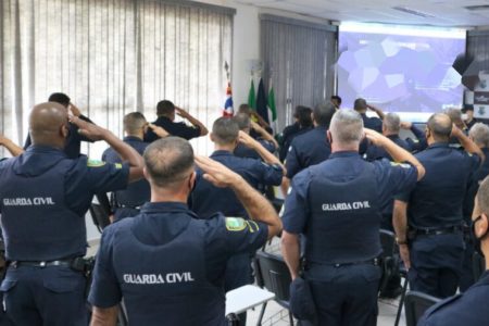 Em foto colorida batalhão de GCMs, de costas, batem continência dentro de sala de reunião - Metrópoles