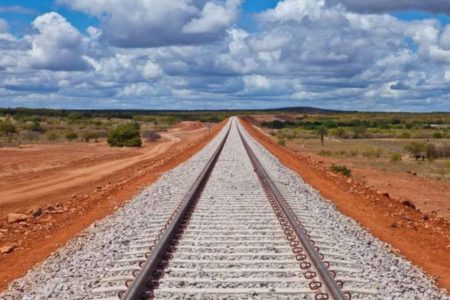 Imagem de trilho de ferrovia cortando uma área de terra e vegetação, com o horizonte ao fundo e céu azul com nuvens - Metrópoles