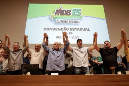 Foto colorida de sete pessoas, com as mãos unidas e os braços para cima; entre elas, há o governador Ibaneis Rocha, ao centro, além dos deputados distritais Wellington Luiz e Hermeto; ao fundo, há uma projeção com informações sobre a convenção estadual do MDB-DF