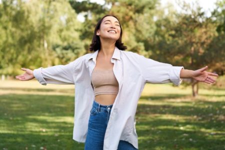 Mulher de blusa branca com braços abertos , olhando para cima com sorriso o rosto no meio da natureza - Metrópoles