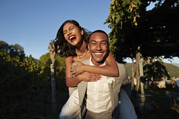 Foto colorida. Mulher está nas costas do homem. Eles estão sorrindo e muito felizes - Metrópoles - hormônios da felicidade