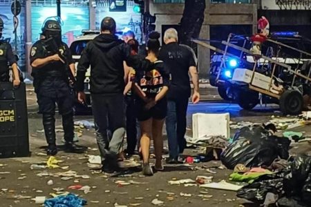 Foto colorida mostra rua cheia de lixo e policiais conduzindo uma mulher suspeita.