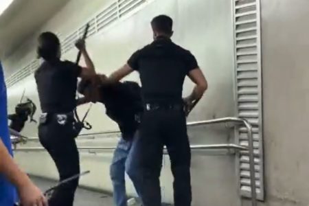 Imagem colorida mostra três funcionários do metrô, de preto, erguendo cacetetes para agredir um homem abaixado tentando se defender