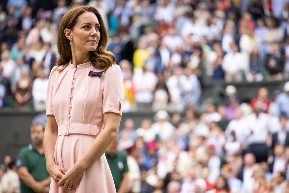 Foto colorida de mulher, branca com cabelos castanhos longos. Ela usa um vestido rosé. Atrás dela, tem uma arquibancada com pessoas - Metrópoles