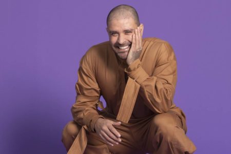 Cantor Diogo Nogueira vestido em macacão caramelo posa sorrindo com uma mão no rosto em fundo roxo - metrópoles