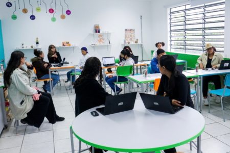 Foto colorida mostra professores e alunos conversando dentro uma sala de aula com paredes e mesas brancas - Metrópoles