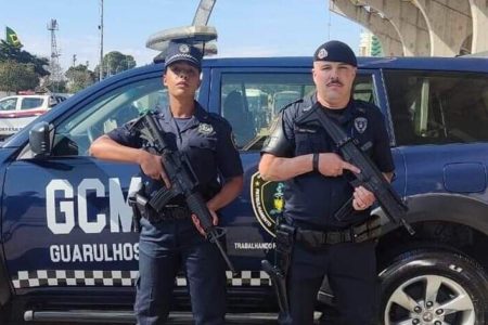 Em foto colorida Guarda Civil Metropolitana feminina segura fuzil, do lado esquerdo da imagem. Ao lado dele um GCM masculino, também empunhando um fuzil. Atrás deles, uma viatura azul escura da GCM de Guarulhos, Grande São Paulo - Metrópoles