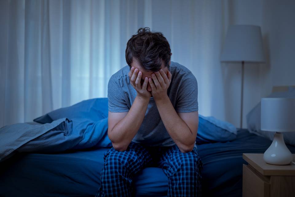 Foto colorida de homem de pijama azul sentado na beira da cama com a mão no rosto e os cotovelos apoiados no joelho - Metrópoles