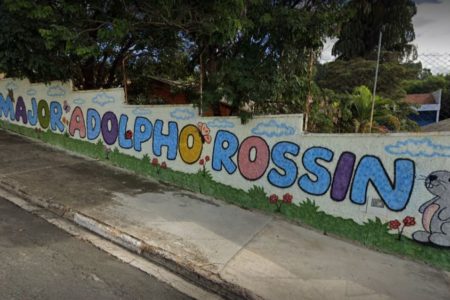 Imagem colorida de fachada de escola com muro pintado com o nome da instituição: "E.E Major Adolpho Rossin", em letras coloridas - Metrópoles