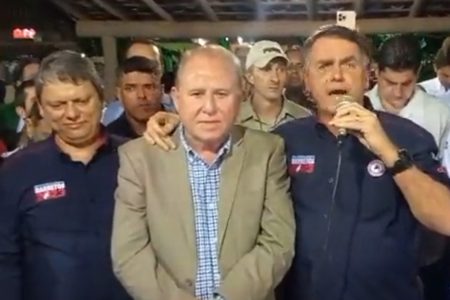 Foto colorida do governador Tarcísio de Freitas à esqueda de camisa azul escura com o ex-presidente Jair Bolsonaro à direita de camisa escura e falando ao microfone - metrópoles