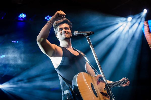 Foto colorida do cantor Luan Santana no palco com um violão pendurado e o pulso cerrado para cima em meios às luzes - Metrópoles