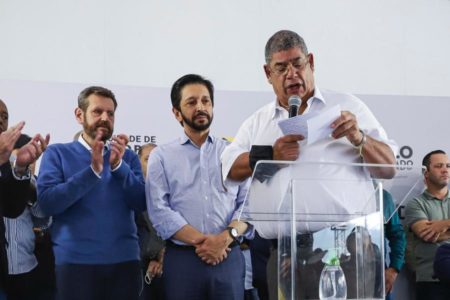 Imagem colorida mostra palco de comício com milton leite, homem negro e grisalho, de camisa branca, lendo um papel no púlpito, no microfone, ao lado de aliados - Metrópoles