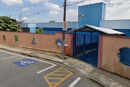 Foto colorida mostra fachada de escola com muro vermelho e portão azul.