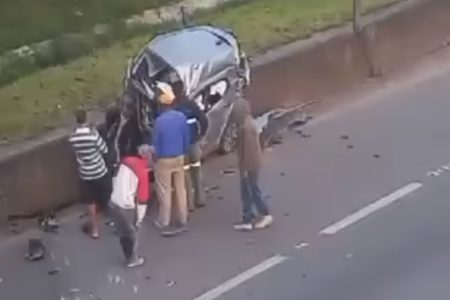 Foto colorida mostra pedestres ao lado das ferragens de veículo alvo de acidente