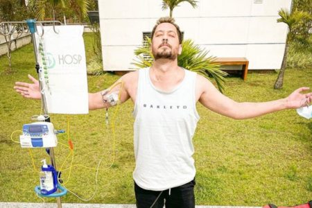 Em foto colorida jovem de camiseta regata branca matém braços abertos, com a cabeça voltada para o céu, de olhos fechados, em frente a um gramado. Ele está com soro no braço - Metrópoles