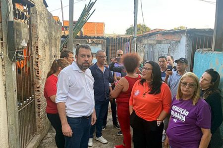 Imagem colorida mostra Guilherme Boulos homem branco de cabelo e barba castanhos, de camisa branca e calça jeans, em meio a um grupo de pessoas em uma viela da comunidade São João, em São Mateus, zona leste de São Paulo - Metrópoles