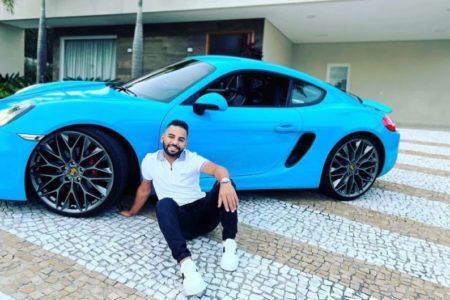 Foto colorida de Felippe Ribeir, da empresa Avanzza, sentado no chão, de camisa branca, calça preta em frente a um carro de luxo azul - metrópoles