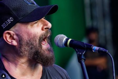 foto colorida mostra o baixista Mingau de camiseta e boné pretos cantando em um microfone - Metropoles