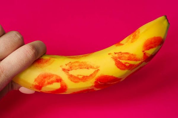 Foto colorida de uma mão feminina segurando uma banana com marcas de batom vermelho em um fundo cor-de-rosa - Metrópoles