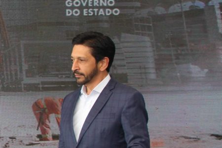 Imagem mostra Ricardo Nunes de terno azul e camisa azul, olhando para sua direita, na frente de um banner do governo do estado, da cintura para cima - Metrópoles