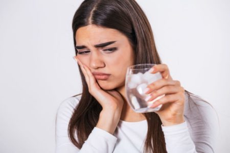 Foto mostra mulher com expressão de desconforto, segurando os dentes enquanto olha um copo de água gelada