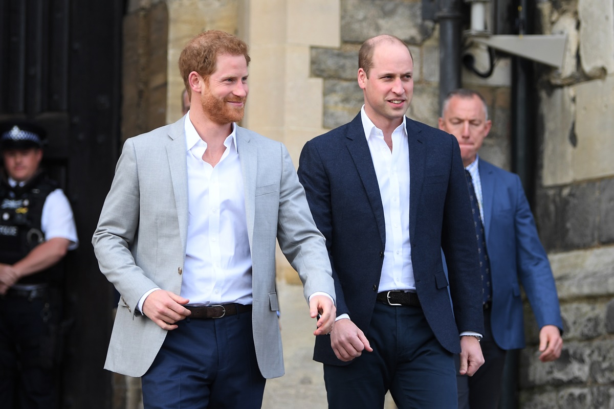 Foto colorida. Doi homens brancos, altos estão andando sorridentes pela rua. Harry, está usando um terno cinza, calça azul escura e uma camisa branca. Já William, está trajando um terno azul escuro e camisa branca - Metrópoles