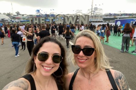 Imagem colorida mostra duas mulheres vestidas de camisetas regadas pretas, de óculos escuros, sorrindo enquanto uma multidão de pessoas anda atrás delas no festival The Town - Metrópoles