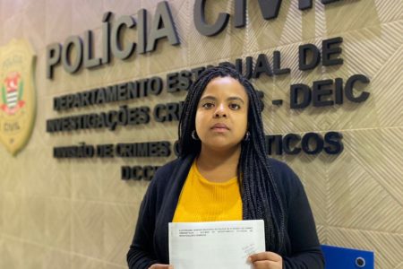 Imagem colorida mostra a vereadora Luana Alves, mulher negra de cabelos pretos, blusa preta e camisa amarela, na frente de uma parede com letreiro da polícia - Metrópoles